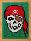 A rectangular knitted blanket featuring a green background and a central skull graphic. The skull wears a red bandana with a white skull and crossbones emblem and a brown eye patch. The design uses black, white, red, and brown yarn.