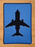 A rectangular chunky knit blanket laid flat on a wooden floor. It features a light blue background, a thick dark navy blue border, and a large black passenger airplane silhouette knit into the center