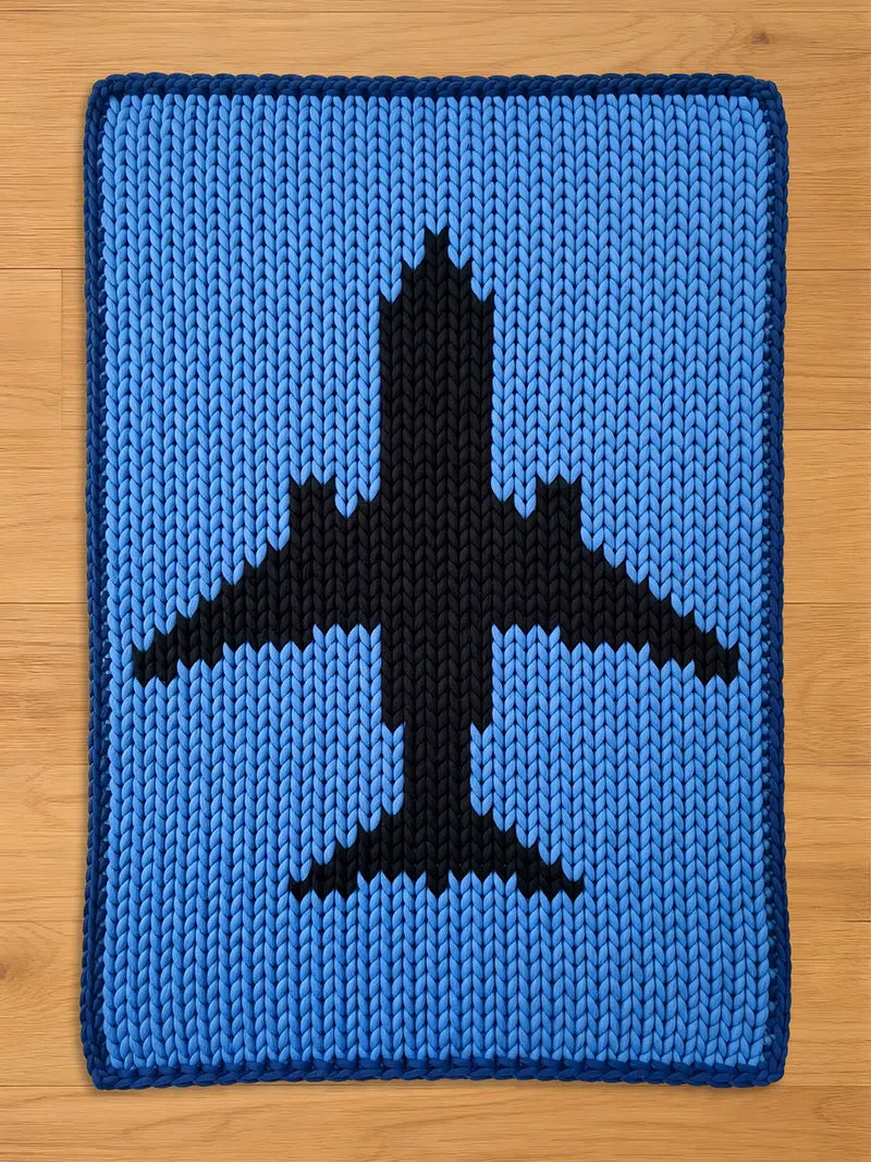 A rectangular chunky knit blanket laid flat on a wooden floor. It features a light blue background, a thick dark navy blue border, and a large black passenger airplane silhouette knit into the center