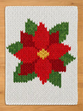 A white chunky knit blanket featuring a large red poinsettia flower pattern with green leaves and a yellow center, displayed flat on a wooden surface.