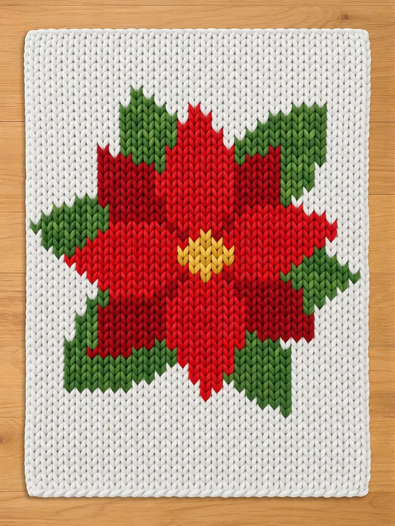 A white chunky knit blanket featuring a large red poinsettia flower pattern with green leaves and a yellow center, displayed flat on a wooden surface.