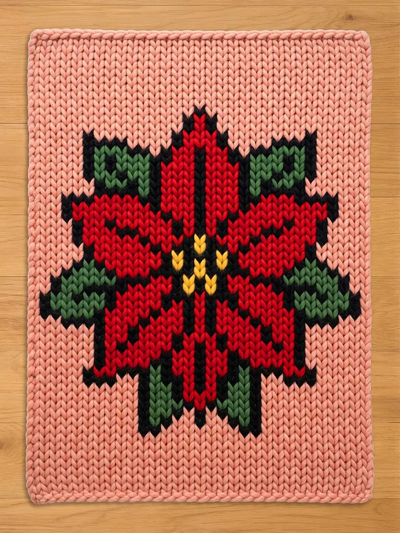 A pink chunky knit blanket featuring a large, stylized red poinsettia flower design in the center. The holiday floral pattern is accented with green leaves, a black border, and yellow details, knitted using thick yarn on a light wood background.