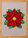 A chunky knit blanket in a light cream or off-white color featuring a large, vibrant red poinsettia flower pattern in the center. The flower has a bright yellow center and is surrounded by green leaves, all knitted in a thick, textured yarn style, displayed flat on a light wood floor