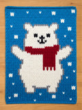 A knitted chunky blanket featuring a white polar bear with a red scarf, set against a blue background with white snowflake accents. The bear has gray accents and a smiling face, creating a cozy, wintery theme.