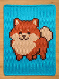 A chunky knit blanket in bright cyan blue featuring a pixel-art style orange and cream Pomeranian dog pattern.