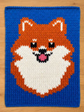 A knitted chunky blanket featuring a Pomeranian dog design. The dog's face is orange, white, black, and pink, set against a vibrant blue background. The knitted texture is clearly visible, adding depth to the design.