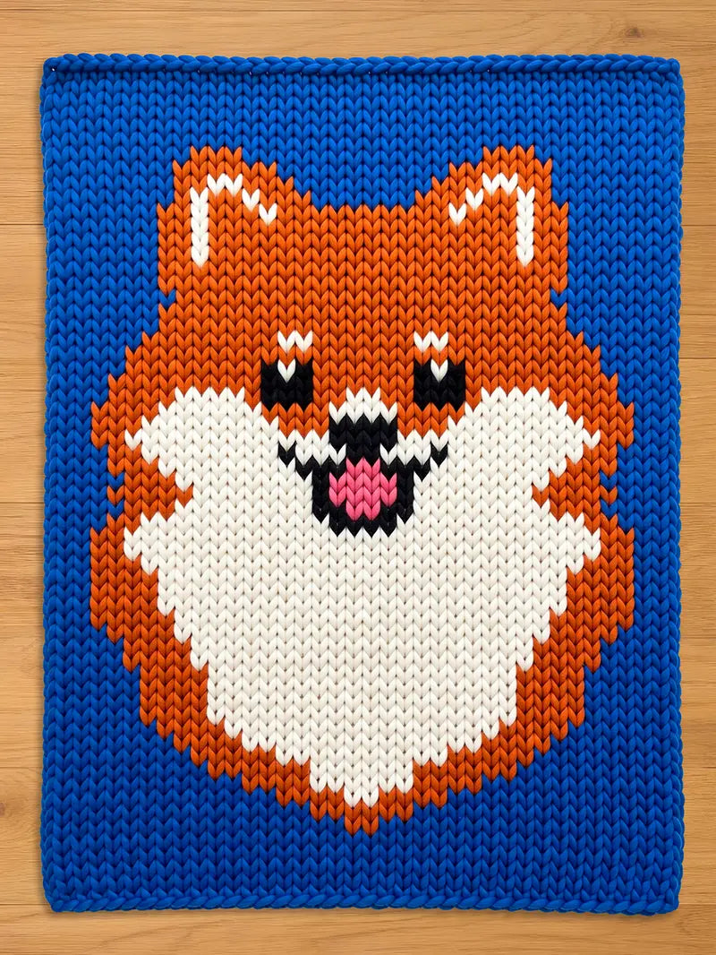 A knitted chunky blanket featuring a Pomeranian dog design. The dog's face is orange, white, black, and pink, set against a vibrant blue background. The knitted texture is clearly visible, adding depth to the design.
