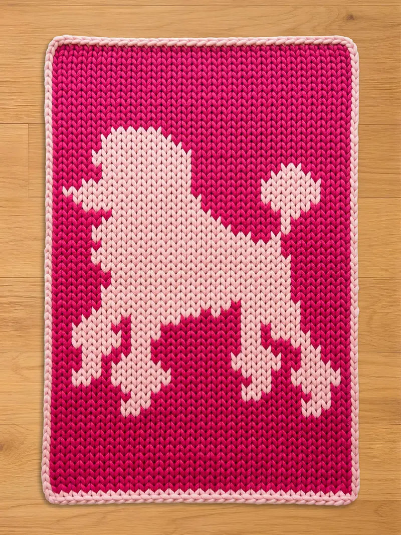 A chunky knit blanket in vibrant magenta pink featuring a light pink knitted poodle silhouette in the center, finished with a light pink border and displayed on a light wood floor.