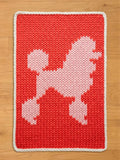 A chunky knit blanket in bright coral red featuring a large, light pink poodle silhouette in the center and a cream white braided border, displayed flat on a light wood floor.