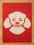 A red knitted chunky blanket with a white dog face design. The dog's face is centered on the rug and features a smiling mouth, closed eyes, and floppy ears. The blanket is rectangular and made of thick yarn.
