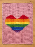 A pink knitted blanket with a large rainbow heart design. The heart features stripes of red, orange, yellow, green, blue, and purple. The blanket is made of thick, chunky yarn.