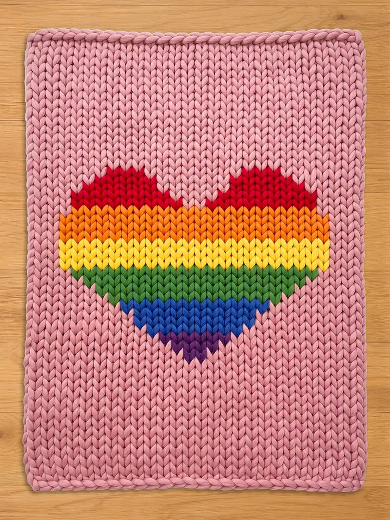 A pink knitted blanket with a large rainbow heart design. The heart features stripes of red, orange, yellow, green, blue, and purple. The blanket is made of thick, chunky yarn.