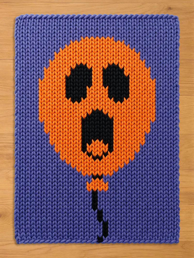 A rectangular knitted blanket featuring a purple background with a large, central orange balloon graphic. The balloon has a black, ghost-like face with two oval eyes and an open, elongated mouth. A thin black string hangs from the balloon.