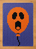 A rectangular knitted blanket featuring a purple background with a large, central orange balloon graphic. The balloon has a black, ghost-like face with two oval eyes and an open, elongated mouth. A thin black string hangs from the balloon.