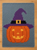A rectangular knitted blanket featuring a gray background with a central orange pumpkin wearing a purple witch hat. The hat includes a black band and a yellow buckle. The pumpkin has a yellow jack-o'-lantern face with eyes and a mouth.