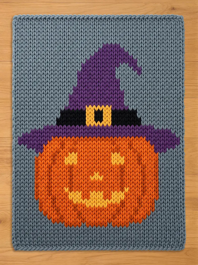 A rectangular knitted blanket featuring a gray background with a central orange pumpkin wearing a purple witch hat. The hat includes a black band and a yellow buckle. The pumpkin has a yellow jack-o'-lantern face with eyes and a mouth.