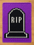 A rectangular knitted blanket featuring a purple border and a central black tombstone motif. The tombstone is outlined in white yarn and displays the white knitted text RIP in the center, with a white base detail at the bottom.