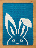 Chunky knit blue blanket featuring a white knitted rabbit pattern with long ears, laid flat on a light wood surface