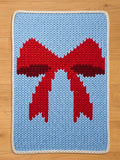 A knitted chunky blanket with a light blue background and a large red bow design. The bow has a darker red center. The rug is bordered with a white knitted trim. The blanket is likely handmade.