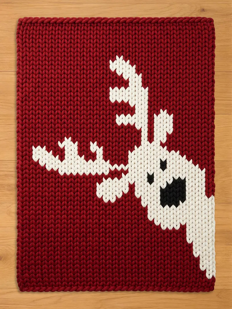 A rectangular chunky knit blanket laid flat on a light wood floor. The blanket is deep red with a pattern of a white reindeer head and large antlers entering from the lower right side, complete with black knit details for the eye and nose
