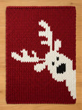 A rectangular chunky knit blanket laid flat on a light wood floor. The blanket is deep red with a pattern of a white reindeer head and large antlers entering from the lower right side, complete with black knit details for the eye and nose