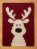 A cozy, handmade-style chunky knit blanket laid flat on a wooden surface. The blanket has a dark red background and showcases a central, festive white reindeer silhouette design with prominent antlers and black facial details.