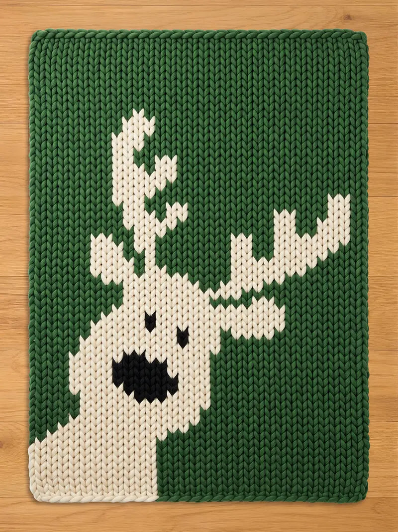 A dark green chunky knit blanket laid flat on a light wooden surface, featuring a large, off-white knitted reindeer silhouette with prominent antlers, black eyes, and a large black nose