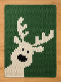 A dark green chunky knit blanket laid flat on a light wooden surface, featuring a large, off-white knitted reindeer silhouette with prominent antlers, black eyes, and a large black nose