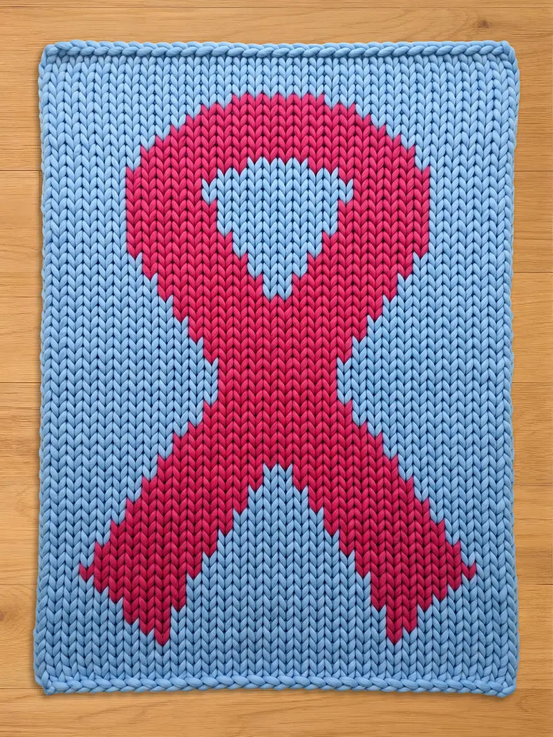 A knitted chunky blanket in light blue with a large pink ribbon design in the center. The ribbon is a symbol of awareness, and the blanket is made of thick, soft yarn. The blanket is displayed on a wooden surface.
