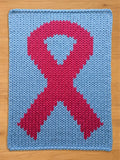 A knitted chunky blanket in light blue with a large pink ribbon design in the center. The ribbon is a symbol of awareness, and the blanket is made of thick, soft yarn. The blanket is displayed on a wooden surface.