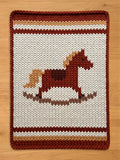 A knitted rectangular chunky blanket featuring a rocking horse design. The rug has a white background with a brown rocking horse in the center. The border and stripes are in shades of brown and tan.
