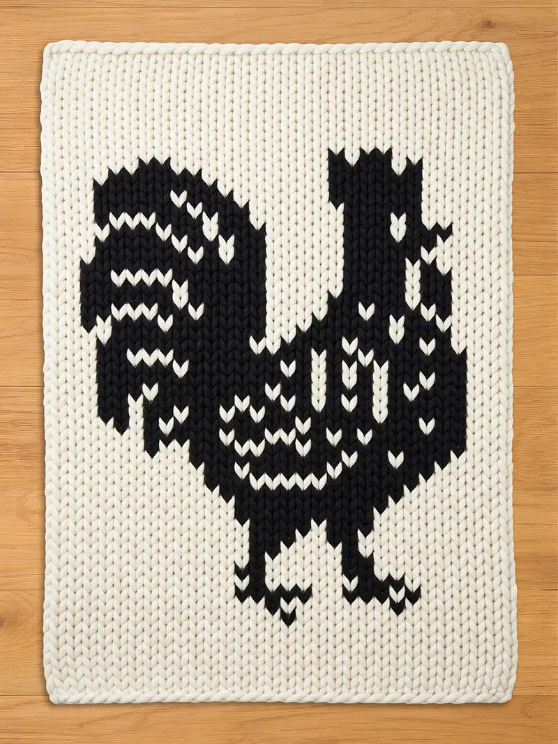 A knitted, off-white chunky blanket featuring a large black rooster design. The rooster is detailed with white accents, creating a striking contrast against the cream background. The blanket is made of thick yarn.