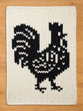 A knitted, off-white chunky blanket featuring a large black rooster design. The rooster is detailed with white accents, creating a striking contrast against the cream background. The blanket is made of thick yarn.