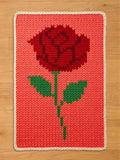 A knitted rectangular blanket featuring a large red rose with green leaves and stem against a coral background. The blanket has a white border and is placed on a wooden surface.