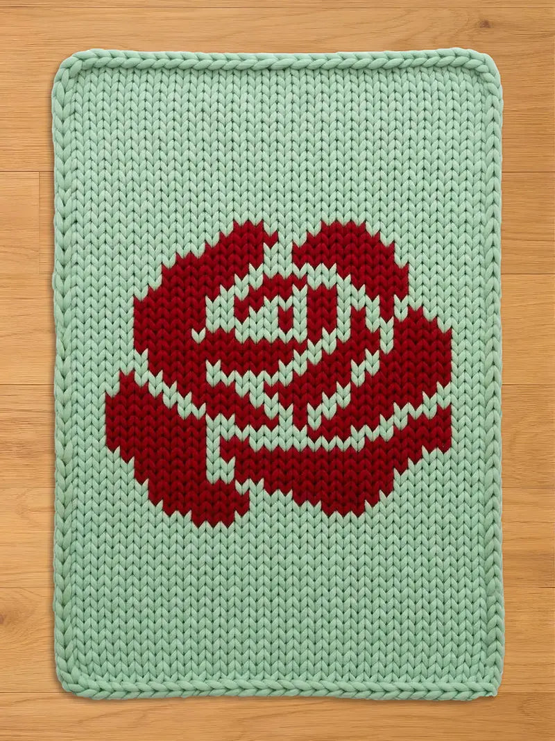 A rectangular, knitted chunky blanket featuring a large red rose design on a light green background. The rose is centrally placed and detailed with white accents. The blanket is made of thick yarn, giving it a textured appearance.