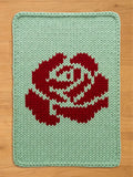 A rectangular, knitted chunky blanket featuring a large red rose design on a light green background. The rose is centrally placed and detailed with white accents. The blanket is made of thick yarn, giving it a textured appearance.
