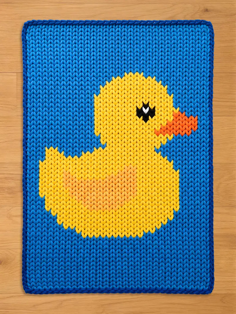 A knitted blue rectangular chunky blanket with a yellow duck design. The duck has an orange beak, black eyes, and a yellow body with an orange center. The blanket has a blue border and is made of a knitted material.