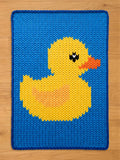 A knitted blue rectangular chunky blanket with a yellow duck design. The duck has an orange beak, black eyes, and a yellow body with an orange center. The blanket has a blue border and is made of a knitted material.