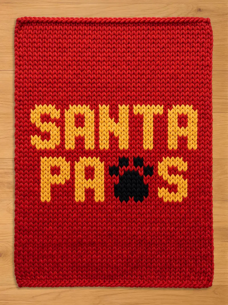 A rectangular red knitted chunky blanket featuring the yellow text SANTA PAWS, with a black paw print replacing the letter W. The knit texture is thick and uniform, creating a bold contrast between the red background and the yellow and black lettering.