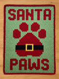 A chunky knitted Christmas-themed blanket with the words "SANTA PAWS" in red letters on a light green background. A red Santa hat-shaped paw print is in the center, with a black belt and yellow buckle. The border is red.