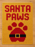 A rectangular yellow knit blanket featuring the red text SANTA PAWS at the top. Below the text is a large red paw print graphic with a black belt and a yellow buckle design integrated into the center of the paw pad.