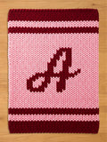 A knitted blanket in shades of pink and burgundy. The blanket features a large letter "A" in burgundy in the center. The top and bottom edges have burgundy stripes, and the rest of the blanket is pink.