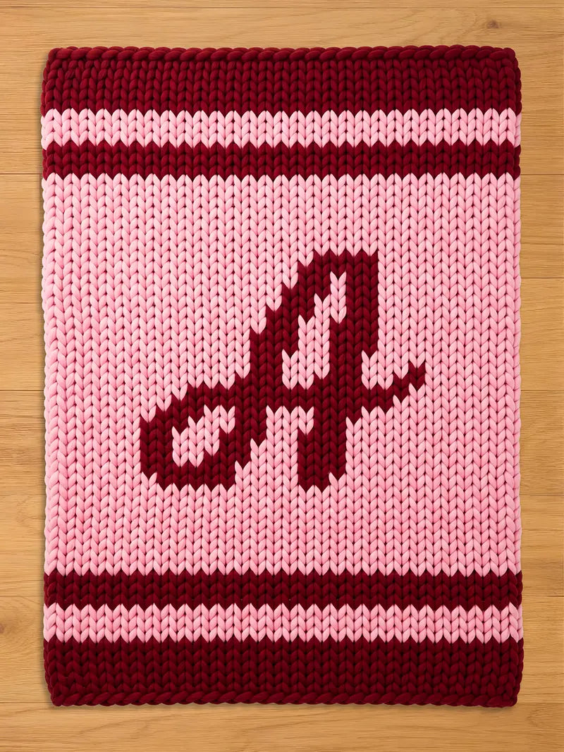 A knitted blanket in shades of pink and burgundy. The blanket features a large letter "A" in burgundy in the center. The top and bottom edges have burgundy stripes, and the rest of the blanket is pink.
