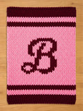 A knitted, rectangular-shaped item with a pink background and a large, dark burgundy letter "B" in the center. The top and bottom edges feature dark burgundy stripes, and the item is made of thick yarn.