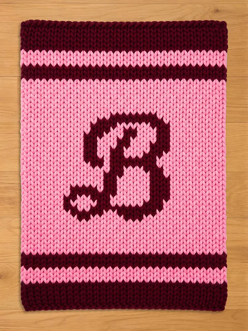 A knitted, rectangular-shaped item with a pink background and a large, dark burgundy letter "B" in the center. The top and bottom edges feature dark burgundy stripes, and the item is made of thick yarn.
