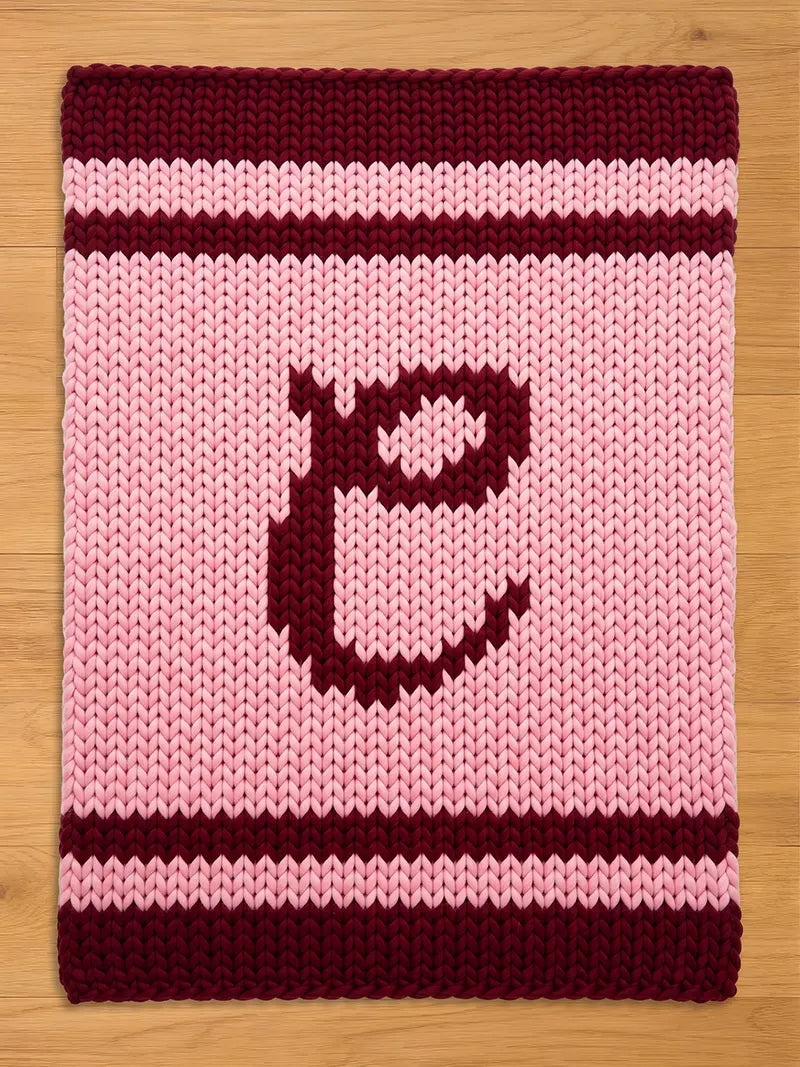 A knitted rectangular blanket in shades of pink and burgundy. The blanket features a large letter "E" in burgundy in the center. Burgundy stripes border the top and bottom edges, with a pink and burgundy striped pattern.