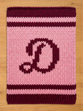 A knitted rectangular chunky blanket in shades of pink and burgundy. The blanket features a large letter "D" in burgundy in the center. The top and bottom edges, along with two stripes, are burgundy. The background is light pink.