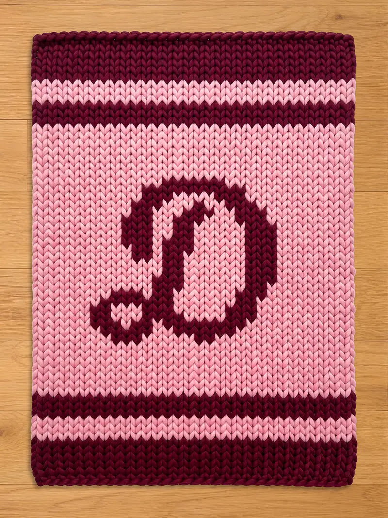A knitted rectangular chunky blanket in shades of pink and burgundy. The blanket features a large letter "D" in burgundy in the center. The top and bottom edges, along with two stripes, are burgundy. The background is light pink.