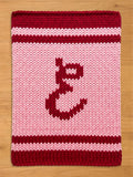 A chunky knitted blanket in shades of pink and burgundy. The blanket features a large, stylized letter "E" in burgundy in the center. The top and bottom edges, along with a stripe, are burgundy. The background is light pink.