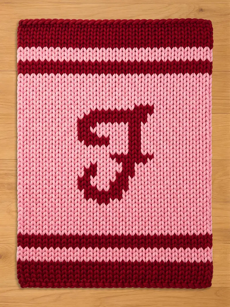 A knitted, rectangular throw blanket in pink and burgundy. The top and bottom borders are burgundy, with a burgundy stripe in the middle. A large burgundy letter "J" is centered on the pink field.